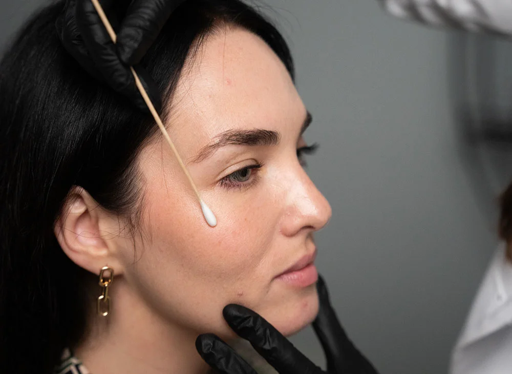 Close up side profile shot of a doctor wearing a lab coat and black plastic gloves swabbing the cheekbone of a younger woman with long black hair. The woman sits calmly while the procedure is performed. - Botox in Aventura, FL