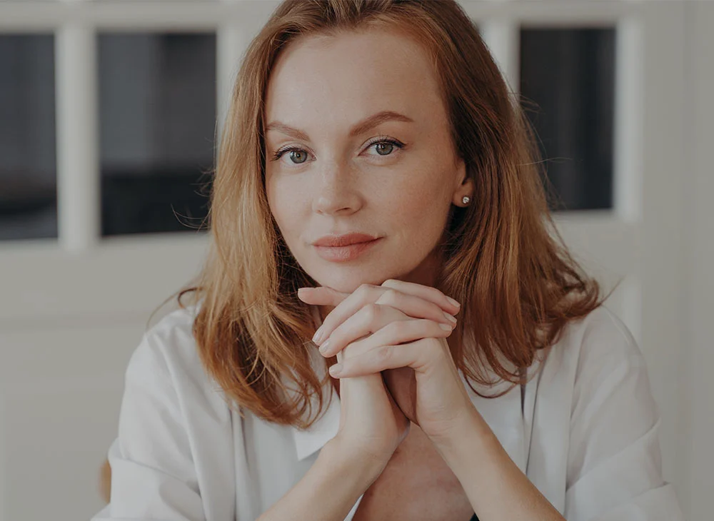 Close-up shot of a red-haired woman resting her head on her folded hands, glancing into the camera with a thoughtful expression. She is wearing a white button-down shirt over a black tank top, exuding a calm and introspective vibe. - Dermal Fillers for Under Eye Bags in Aventura, FL