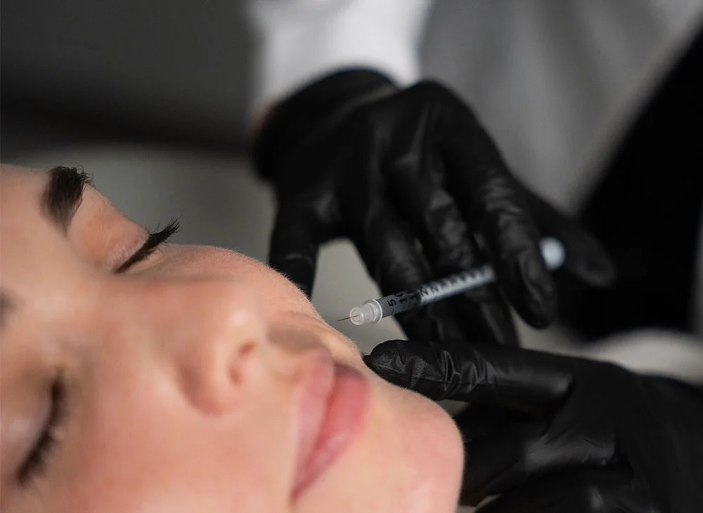 Close-up shot of a woman receiving cheek dermal fillers, with a doctor wearing black latex gloves carefully administering the injection. The scene emphasizes precision and professionalism. - Dermal Fillers in Aventura, FL
