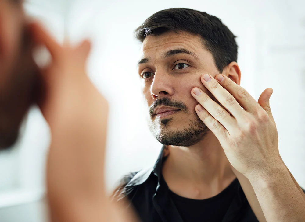 Close-up shot of a man looking at himself in a mirror, gently touching his cheek. He has short black hair, facial hair, and is wearing a black undershirt layered with a black over shirt. - Ectropion Repair Surgery in Aventura, FL