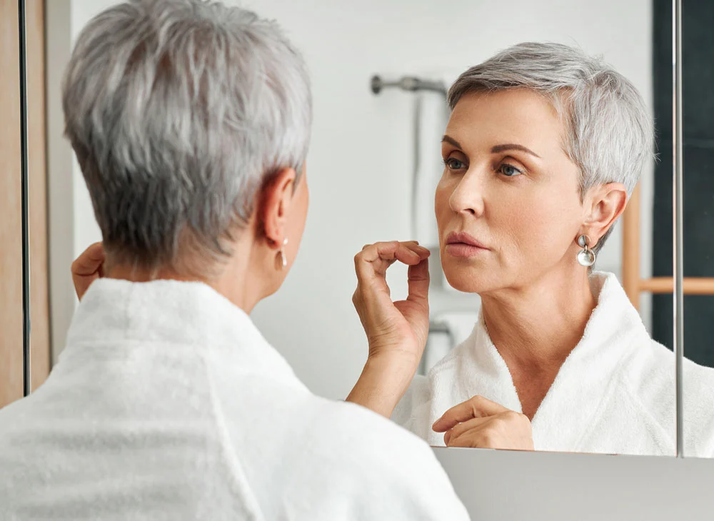 Close up shot from the back of a middle-aged woman with short grey hair, wearing a white robe, looking into a mirror. She holds one hand up, framing the side of her face as she examines herself. - Facetite Accutite in Aventura, FL