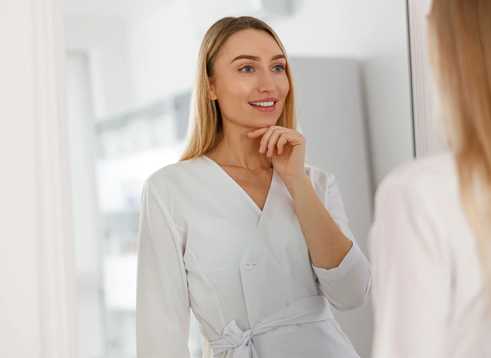 A woman with long blonde hair wearing a white robe, smiling as she stands in front of a mirror. She holds her hand on her chin, and the background is softly blurred, creating a serene and reflective atmosphere. - Facial Fat Grafting in Aventura, FL
