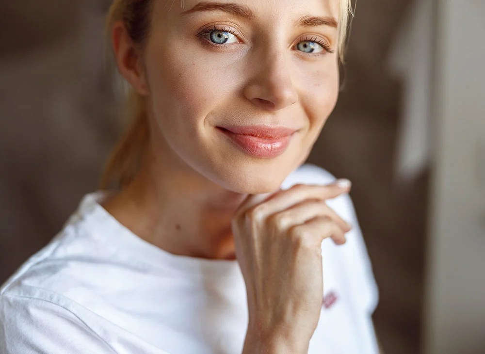 Close-up shot of a young blonde woman with her hair styled in a ponytail. She has one hand held under her chin and is looking at the camera with a soft, closed-mouth smile. She is wearing a white T-shirt, exuding a casual and approachable vibe. - Hand Rejuvenation in Aventura, FL