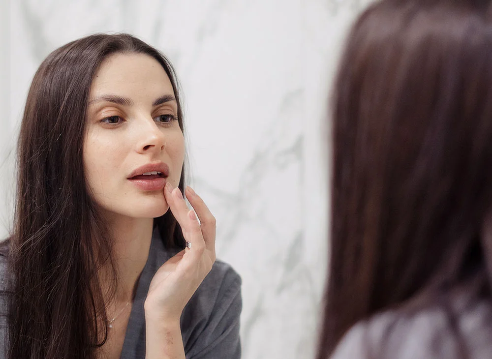 A woman with long dark hair looks into a mirror while gently touching her lip with one hand. She is wearing a grey shirt, appearing thoughtful and focused. - Lip Injections in Aventura, FL