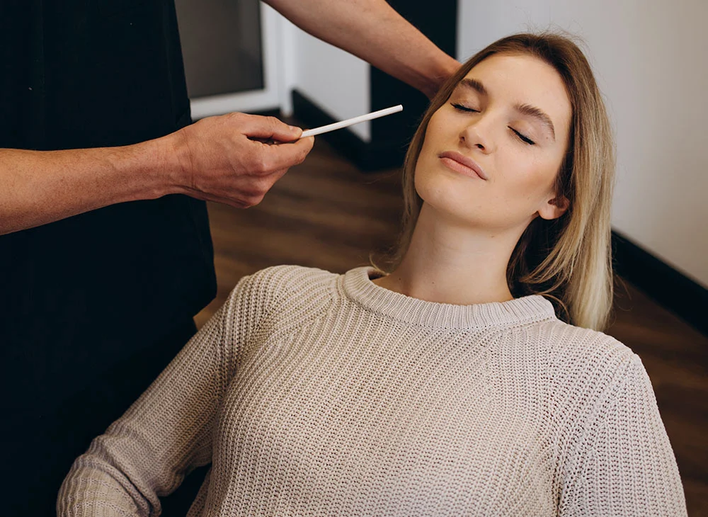 A woman with blonde hair, wearing a cream-colored sweater, lies on a consulting room chair with her eyes closed. The doctor examines her face with a white stick, performing an assessment or consultation. - Lump Bump Eyelid in Aventura, FL