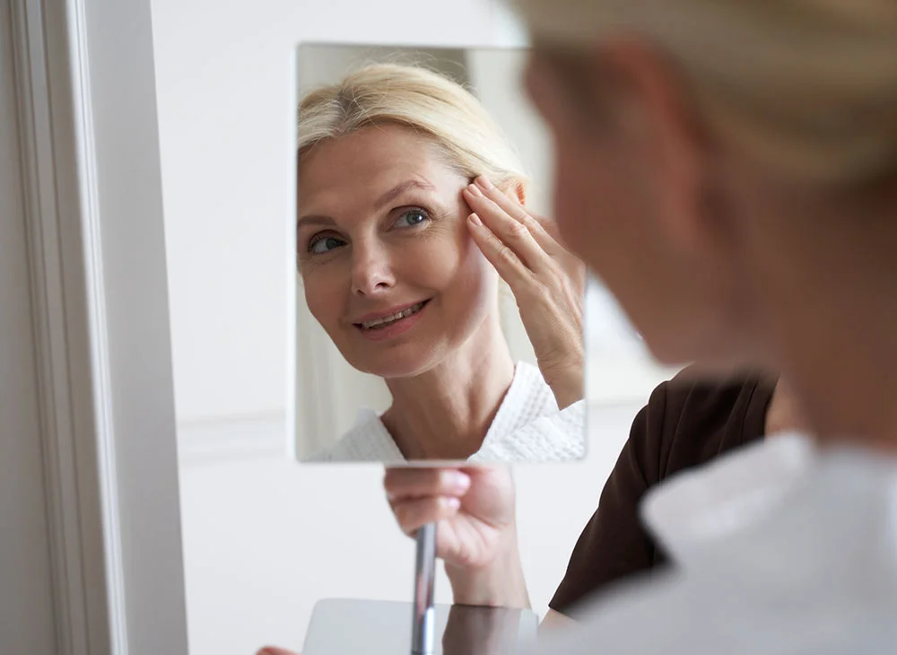 A middle-aged woman with blonde hair up in a bun smiles into a mirror held by a nurse. She is smiling while gently grazing her cheek with one hand, wearing a white robe, radiating a sense of satisfaction and joy. - Mid Facelift in Aventura, FL
