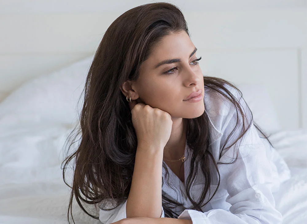 A woman in a white shirt sits on a bed, resting her head on her hand. With long brown hair, she looks to the side, gazing past the camera in a thoughtful or contemplative manner. - Non-Surgical Rhinoplasty in Aventura, FL