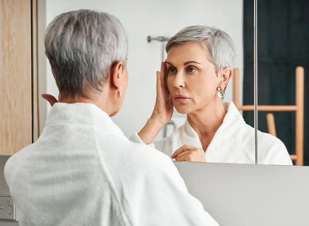 Shot from the back of a middle-aged woman with short grey hair, wearing a white robe, looking into a mirror. She holds one hand up, framing the side of her face as she examines herself. - Scar Revision in Aventura, FL