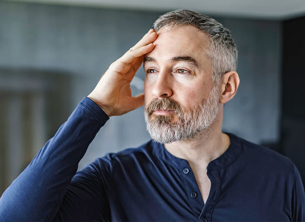 A portfolio shot of a man facing slightly to the side of the camera, with one hand resting on his head as he gazes outward. He is wearing a blue long-sleeve shirt and has white short hair and a beard. - Surgery for Tearing Disorder in Aventura, FL