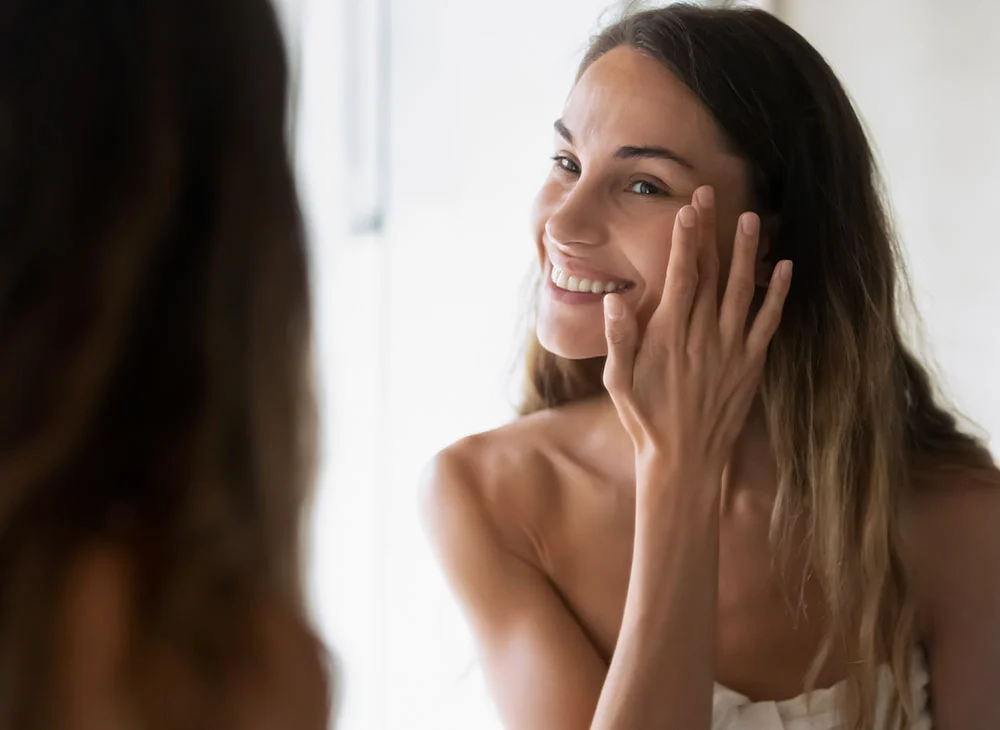A woman with long brown hair and blonde highlights looks in the mirror, smiling as she touches her under-eye area with one hand. She is wearing a towel, radiating a relaxed and confident demeanor. - Xanthelasma in Aventura, FL